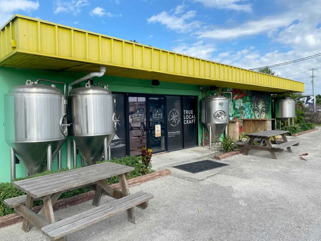 1981 Brewing Co. exterior with brewing tanks, patio, picnic tables, and vibrant mural. Cayman Islands.