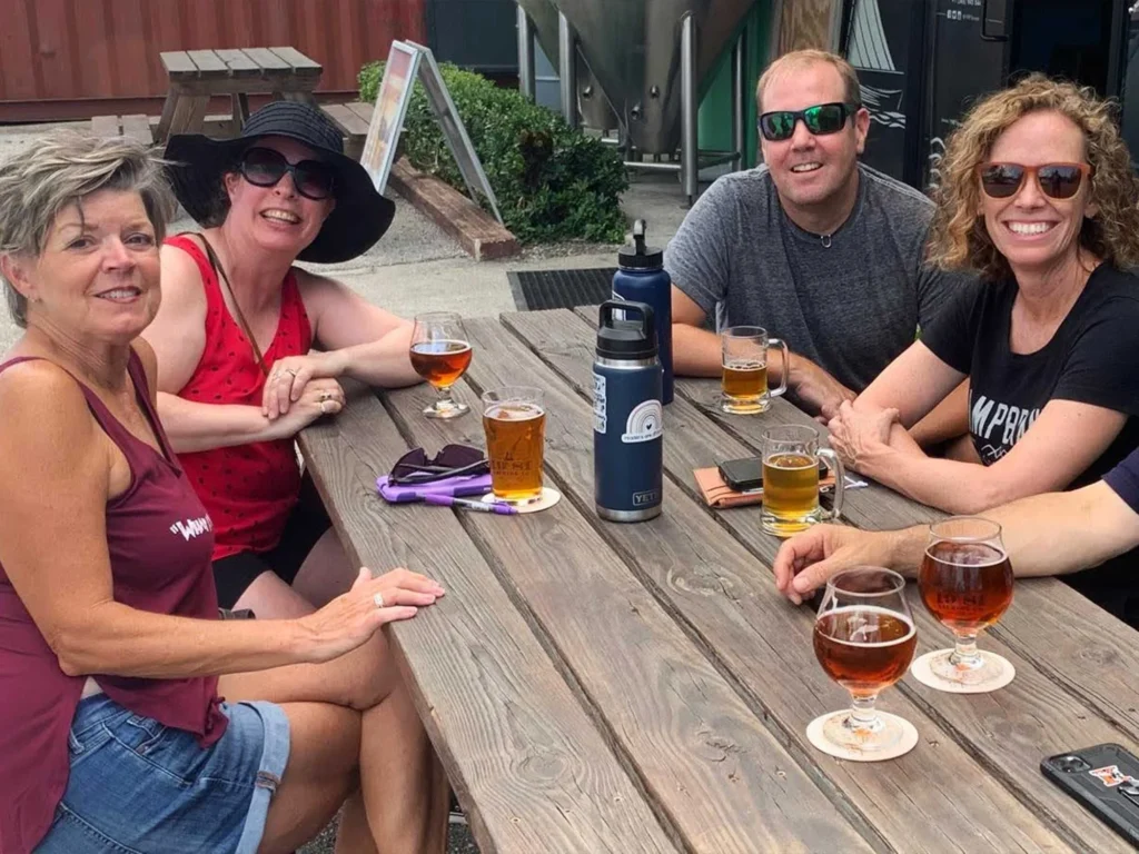 Four friends enjoy craft beer at an outdoor brewery picnic table.