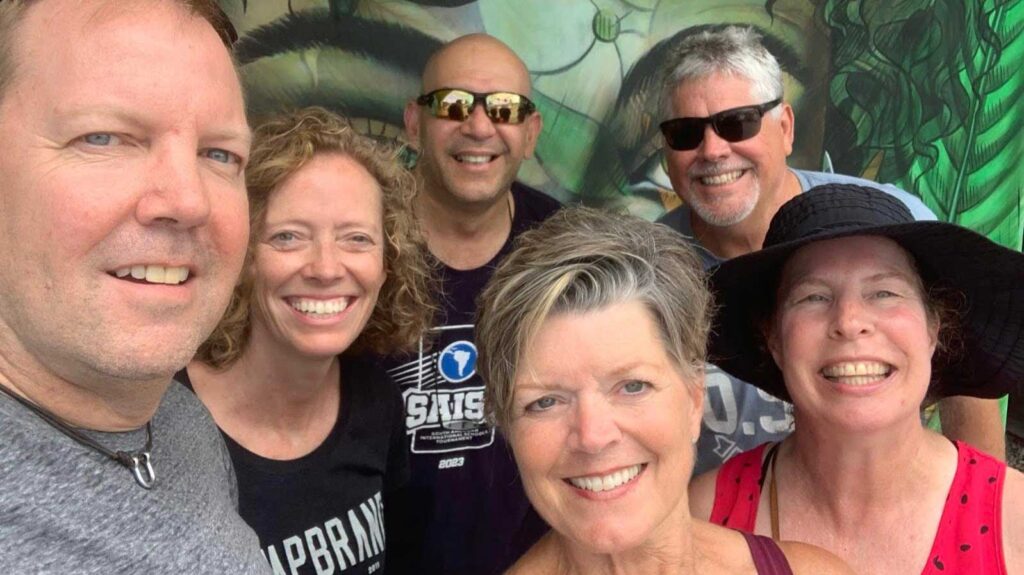 Six friends smile for a selfie in front of a colorful mural.
