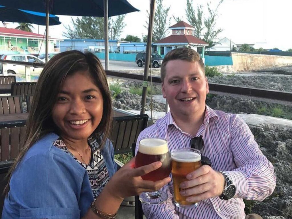 Two people smiling and holding glasses of 19-81 beer at an outdoor restaurant in the Cayman Islands