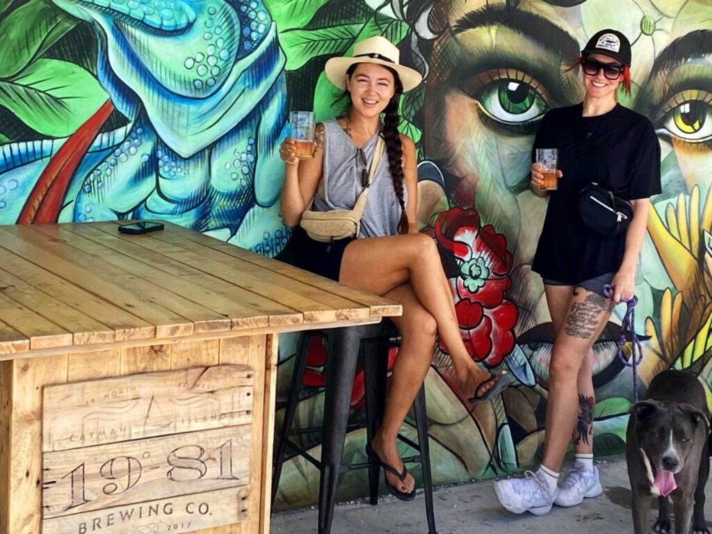 Two women with a dog, smiling and drinking 19-81 Cayman beer in front of the mural at the 19-81 Brewing Co. taproom