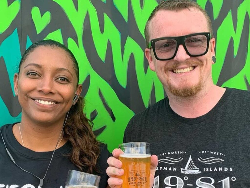 A couple smiling and drinking beer in front of the mural at the 19-81 Brewing Co. taproom.