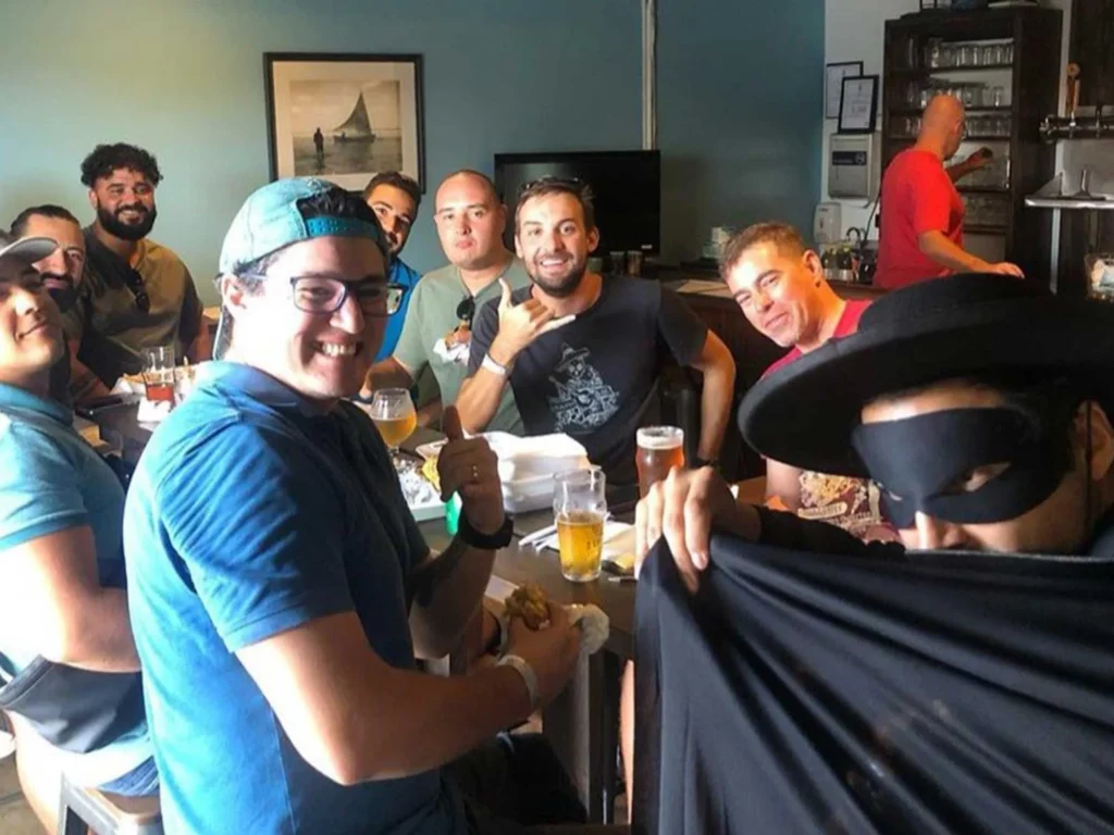Friends enjoy beers and food at a taproom; one wears a Zorro-style mask.